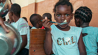 Ein Mädchen sitzt zwischen anderen Kindern und isst mit einem Löffel Porridge