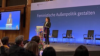 Ministerin Baerbock steht auf einer Bühne und spricht. Im Hintergrund steht der Schriftzug "Feministische Außenpolitik"