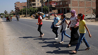 Eine Gruppe Mädchen läuft mit einer Frau über eine Straße. Sie halten sich gegenseitig an der Hand, von links kommen Motorräder auf sie zu.