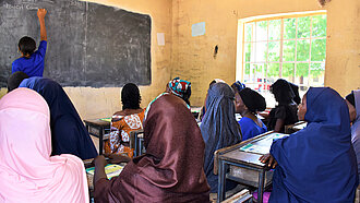 Ansicht einer Schulklasse in Nigeria, die Schülerinnen sind von hinten zu sehen und blicken auf die Tafel