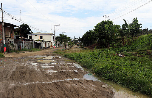 Eine matschige Straße, rechts daneben grüne Gräser und Palmen, links kleine Häuser beziehungsweise Hütten 