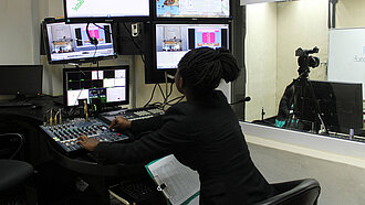 Eine Frau sitzt vor Monitoren und einem Schaltpult.