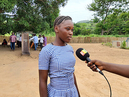 Adèle, ein Mädchen aus Guinea, steht mir ihrer Schuluniform vor einem Mikro und spricht hinein. Im Hintergrund sind Mitglieder der Schule zu erkennen.