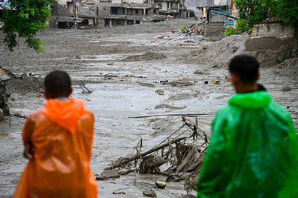 Zwei Personen in bunten Regenponchos (orange und grün) stehen am Bildrand und blicken auf eine überschwemmte, schlammige Straße. Im Hintergrund sind stark beschädigte Gebäude zu sehen. Die Umgebung wirkt verwüstet und es liegen Trümmer sowie Gegenstände verstreut.