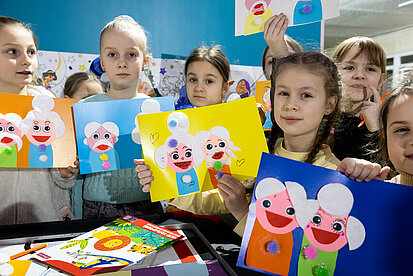 Eine Gruppe von Mädchen zeigt stolz ihre farbenfrohen Kunstwerke in einem kinderfreundlichen Raum. Jedes Mädchen hält ein selbstgemaltes Bild mit Figuren, die aus Pompons und Papier bestehen. Die Mädchen lächeln, während sie ihre Kunstwerke präsentieren.