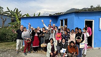 Gruppenfoto einer Projektreise in Nepal, Unterstützende und Projektteilnehmende mit einem blauen Gebäude und dem Himalaya im Hintergrund.