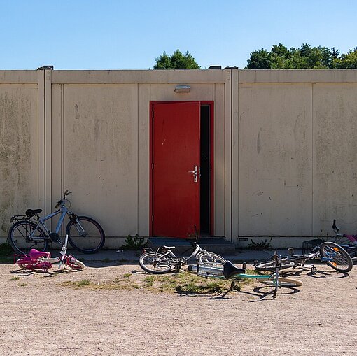 Eine leicht geöffnete rote Tür in einem Container, davor stehen und liegen einige Fahrräder