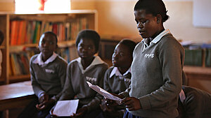 Eine Schülerin in Schuluniform liest vor einer Gruppe von Mitschülerinnen in einem Klassenzimmer in Malawi vor. Im Hintergrund sind Bücherregale zu sehen.