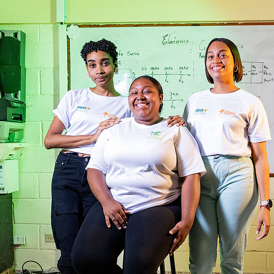 Drei junge Frauen stehen und sitzen gemeinsam in einem Schulungsraum; eine Person sitzt auf einem Hocker, zwei weitere stehen unterstützend daneben, im Hintergrund eine Tafel mit technischen Zeichnungen und Geräteausstattung.