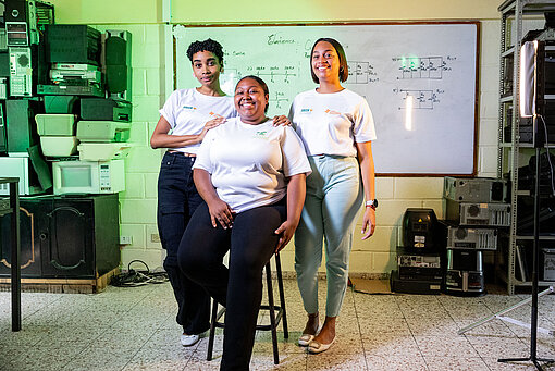 Drei junge Frauen stehen und sitzen gemeinsam in einem Schulungsraum; eine Person sitzt auf einem Hocker, zwei weitere stehen unterstützend daneben, im Hintergrund eine Tafel mit technischen Zeichnungen und Geräteausstattung.