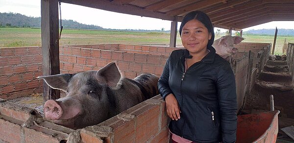 Eine junge Frau steht in einem überdachten Stall neben mehreren Schweinen, die in gemauerten Buchten stehen; hinter ihr sind Felder zu sehen.