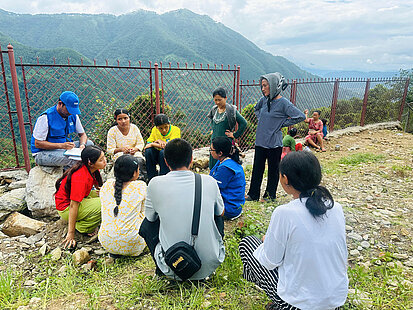 Plan International Mitarbeitende sprechen mit betroffenen jungen Frauen ch einer Naturkatastrophe in Nepal, umgeben von Bergen und einem Zaun.