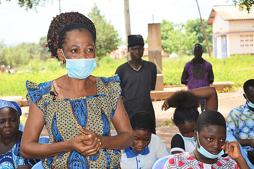 Eine Frau mit Mund-und-Nasenschutz spricht vor einer Gruppe Menschen.