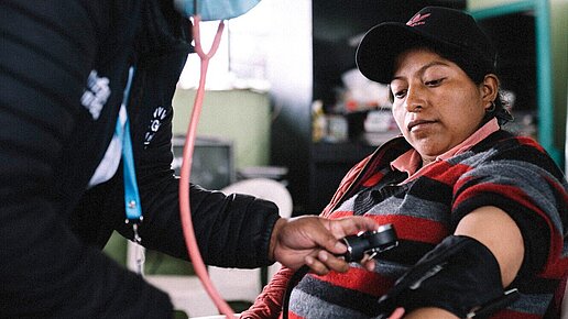 Eine Frau Sitzt auf einem Stuhl und bekommt ihren Blutdruck gemessen.