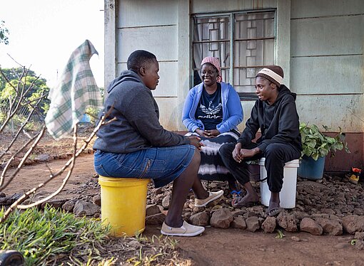Zwei Frauen und ein Mann sitzen zusammen vor einem Haus auf Hockern und unterhalten sich