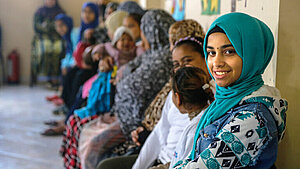 Junges Mädchen mit türkisfarbenem Kopftuch lächelt in die Kamera, im Hintergrund sitzen weitere Frauen und Mädchen in einer Beratungseinrichtung in Ägypten.