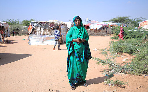Eine Frau in grünem Gewand steht vor einem Camp. Sie hat die Hände ineinander verschränkt und schaut in die Kamera
