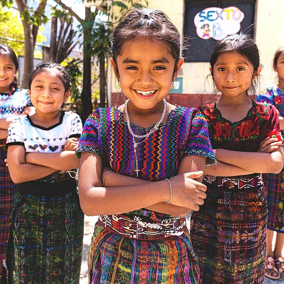 Eine Gruppe von Mädchen aus Guatemala mit bunten Kleidern steht in einem Dreieck. Alle haben ihre Arme vor der Brust verschränkt und lächeln entschlossen.