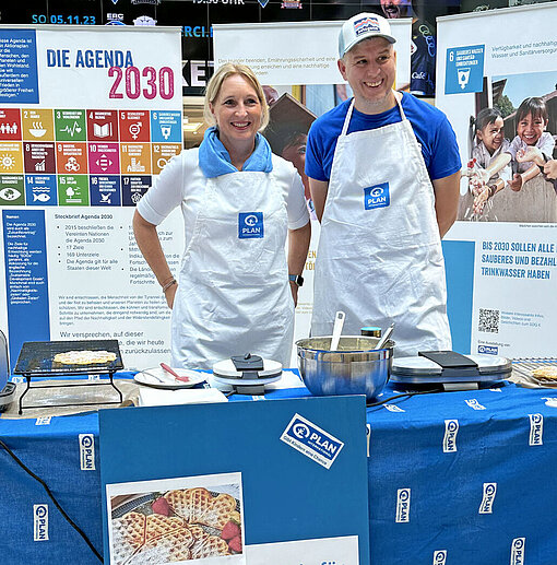 Eine Frau und ein Mann mit Schürze backen Waffeln vor einer Info-Wand mit UN-Nachhaltigkeitszielen