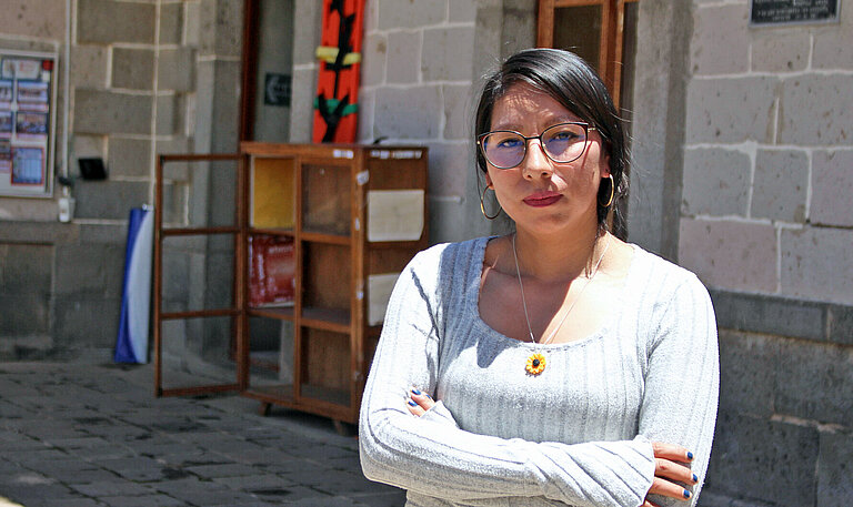 Eine junge Frau in weißem Pullover und mit Brille steht selbstbewusst mit verschränkiten Armen vor einem Gebäude