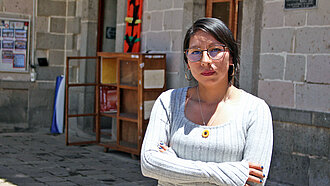 Eine junge Frau in weißem Pullover und mit Brille steht selbstbewusst mit verschränkiten Armen vor einem Gebäude