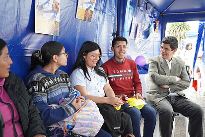 Eine gemischte Gruppe aus Jugendlichen und Erwachsenen, darunter junge Frauen und Männer, sitzt entspannt auf weißen Kunststoffstühlen in einem Zelt. Sie unterhalten sich, einige lächeln. An den blauen Zeltwänden hängen bunte Poster.