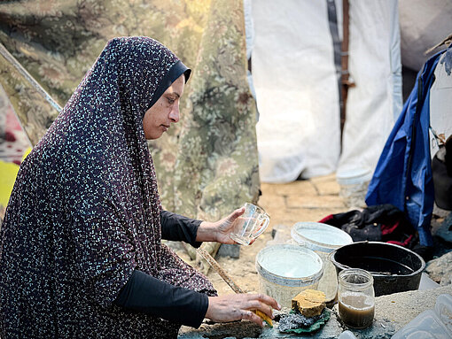 Eine Mutter aus Gaza bereitet im Freien Essen zu. Um sie herum sind Zelte und Schutt.