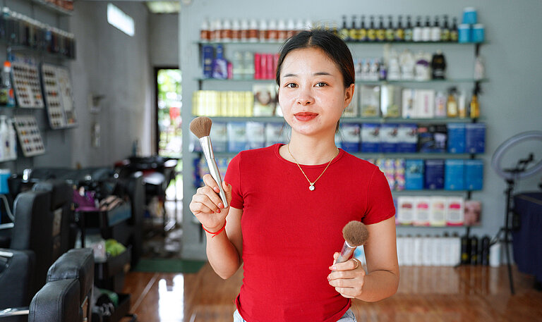 Eine junge Asiatin in rotem T-Shirt steht inmitten eines Friseursalons und schaut in die Kamera