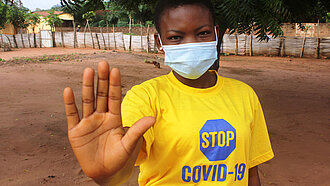Ein Mädchen, auf dessen Tshirt steht "Stop Covid-19", schaut mit Mund-Nasen-Schutz in die Kamera und macht mit der Hand eine abwehrende Haltung