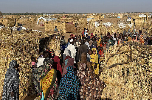 Menschen laufen durch ein Camp für Geflüchtete in Sudan, es stehen Hütten aus Stroh im Camp