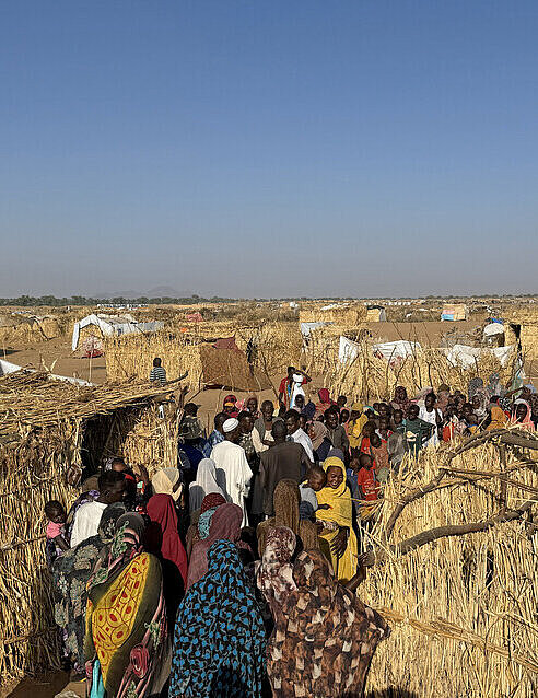 Menschen laufen durch ein Camp für Geflüchtete in Sudan, es stehen Hütten aus Stroh im Camp