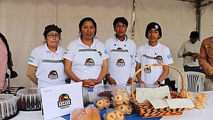 Vier Personen präsentieren Gebäck an einem Verkaufsstand der Bäckerei „Espiga Dorada“ in Ecuador. Auf dem Tisch liegen Kuchen, Brot und Kekse.