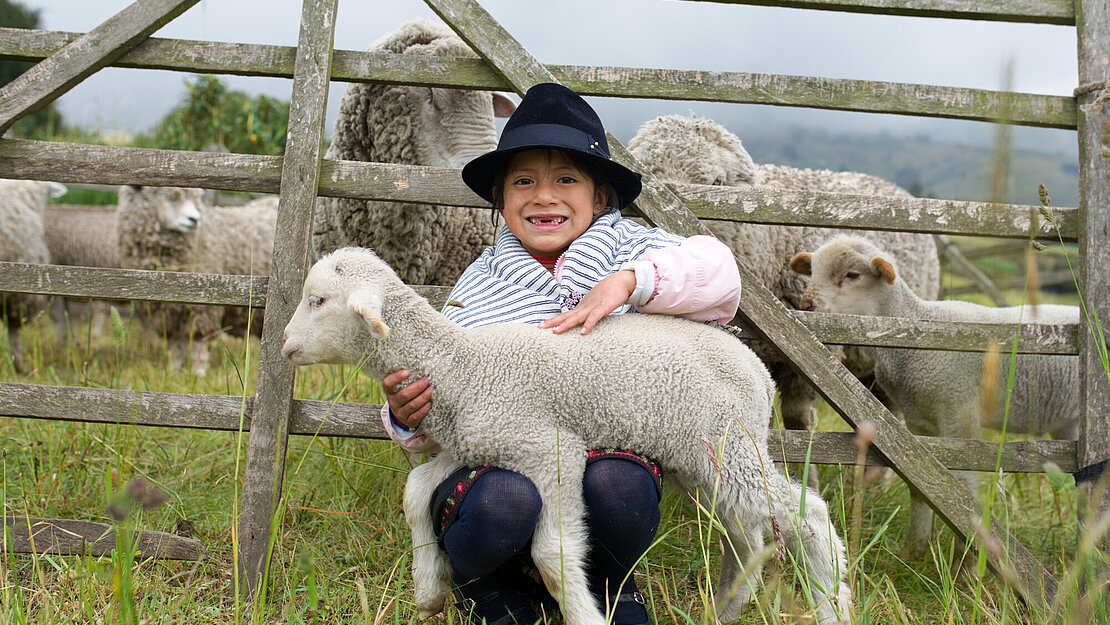 Ein Mädchen sitzt vor einem Weidezaun und hat ein Lamm auf dem Schoß