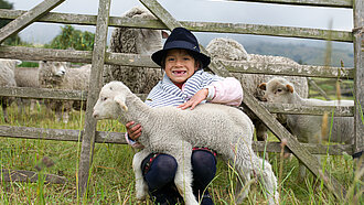 Ein Mädchen sitzt im Gras vor einem Weidezaun und hält ein Lamm im Arm