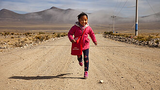Fröhliches Mädchen läuft lachend auf einer staubigen Straße im bolivianischen Hochland, mit Bergen und Nebel im Hintergrund.