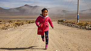 Lachendes Mädchen in pinker Jacke rennt auf einer Straße im bolivianischen Hochland Richtung Kamera. Fröhliches Mädchen läuft lachend auf einer staubigen Straße im bolivianischen Hochland, mit Bergen und Nebel im Hintergrund.