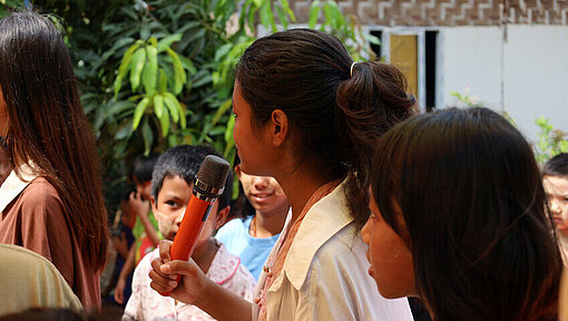 Nan (Mitte) engagiert sich als Freiwillige in einem der Kinderschutzräume in Mandalay Eine junge Frau steht mit einem Handmikrofon in einer Gruppe anderer junger Frauen