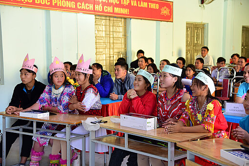 Viele Jugendliche sitzen in einem Klassenzimmer an Tischen, in der vorderen Reihe tragen sechs Jugendliche Papier-Kronen, von denen jeweils drei Jugendliche die gleiche tragen