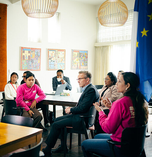Mehrere Menschen, darunter ein Mädchen im pinken Pullover, sitzen an einem runden Tisch und folgen einer Präsentation