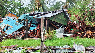 ein zerstörtes, auf die Seite gekipptes Haus umrahmt von abgeknickten Palmen