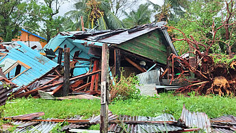 ein zerstörtes, auf die Seite gekipptes Haus umrahmt von abgeknickten Palmen