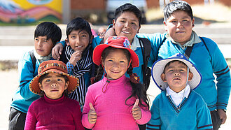Eine Gruppe von Kindern aus Bolivien guckt glücklich in die Kamera, im Vordergrund steht ein Mädchen mit pinkem Pullover, sie zeigt zwei Daumen nach oben.