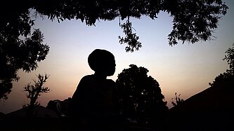 Eine Person steht in der Natur bei aufgehender Sonne. Die Umgebung und die Person sind schattiert, nur der Himmel leuchtet farbig und erzeugt eine melancholische Stimmung.