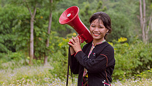 Junge Frau aus Vietnam hält ein Megaphon in einem ländlichen Umfeld und lächelt.