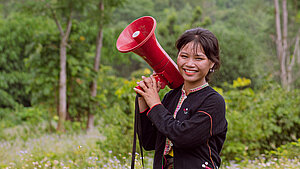 Junge Frau aus Vietnam hält ein Megaphon in einem ländlichen Umfeld und lächelt.