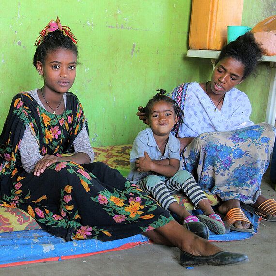 Eine Mutter sitzt mit zwei Kindern auf einer Matratze vor einer grünen Wand. Alle drei blicken in die Kamera; das jüngste Kind lächelt. Neben ihnen stehen Wasserkanister und Haushaltsgegenstände.