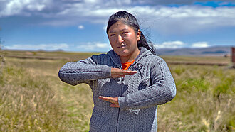 Geovana, eine junge Frau, steht in einem Feld und macht mit ihren Armen ein Gleichzeichen. 