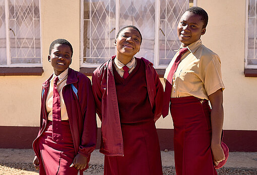 Drei Mädchen in rot-weißer Schuluniform stehen vor einer Wand und lächeln in die Kamera