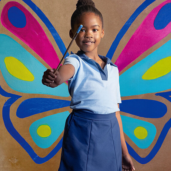 Ein lächelndes Mädchen steht vor einer Wand mit bunten, gemalten Schmetterlingsflügeln und hält einen Pinsel nach vorne ausgestreckt.