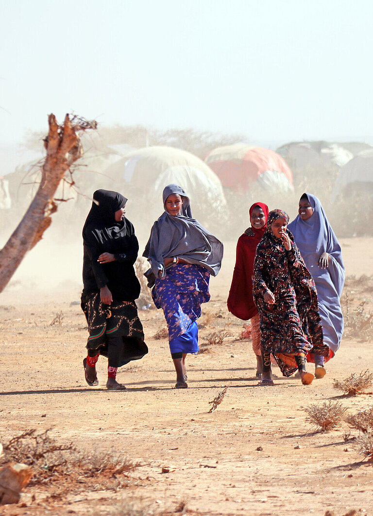 Eine Gruppe heranwachsender Mädchen läuft durch eine trockene Landschaft, im Hintergrund ist ein Camp mit vielen Zelten zu erkennen
